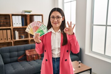 Wall Mural - Young latin woman working at consultation office holding money doing ok sign with fingers, smiling friendly gesturing excellent symbol