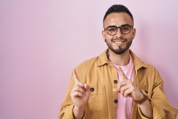 Poster - Young hispanic man standing over pink background smiling and looking at the camera pointing with two hands and fingers to the side.