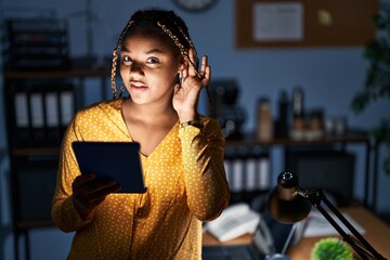 Wall Mural - African american woman with braids working at the office at night with tablet smiling with hand over ear listening an hearing to rumor or gossip. deafness concept.