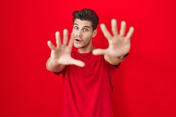 Sticker - Young hispanic man standing over red background doing stop gesture with hands palms, angry and frustration expression