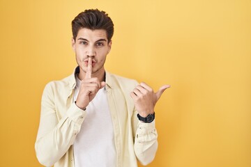 Sticker - Young hispanic man standing over yellow background asking to be quiet with finger on lips pointing with hand to the side. silence and secret concept.