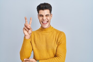 Sticker - Young hispanic man standing over blue background smiling with happy face winking at the camera doing victory sign with fingers. number two.