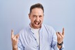 Middle age caucasian man standing over blue background shouting with crazy expression doing rock symbol with hands up. music star. heavy music concept.