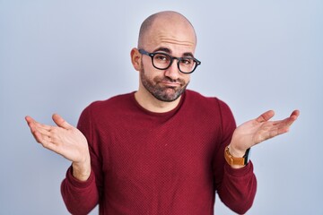 Wall Mural - Young bald man with beard standing over white background wearing glasses clueless and confused expression with arms and hands raised. doubt concept.