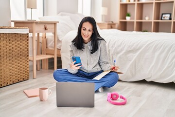 Wall Mural - Young caucasian woman student writing on book using smartphone at bedroom