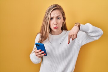 Sticker - Young blonde woman using smartphone typing message pointing down looking sad and upset, indicating direction with fingers, unhappy and depressed.