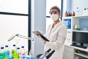 Poster - Young blonde woman wearing scientist uniform and medical mask writing on clipboard at laboratory