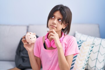 Poster - Young girl holding piggy bank serious face thinking about question with hand on chin, thoughtful about confusing idea