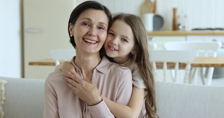 Wall Mural - Mature 50s grandmother and cute grandkid child enjoying leisure together on home couch, looking at camera, laughing, feeling joy, cheerful, happy. Kid hugging grandma with love, affection