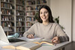 © fizkes - Portrait of pretty teen girl student studying in library, doing homework sit at desk smile staring at camera enjoy studies, gains new knowledge, practicing language, prepare for university admission