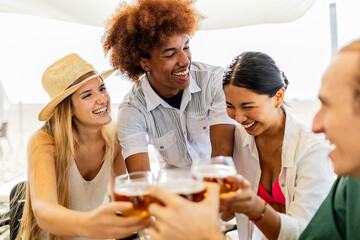 Smiling young ethnic group of millennial friends cheering with beer at beach bar. Multi-ethnic happy people enjoying celebrating together on summer vacation.