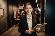 © aicandy - Portrait of a stunning event planner with a warm smile, wearing a chic blazer and holding a clipboard while standing in front of an elegant event space, generative ai