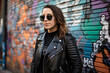 © aicandy - Portrait of a stylish social media manager with a cool and confident vibe, wearing a leather jacket and sunglasses while posing against a graffiti wall, generative ai