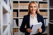 © aicandy - Smiling insurance agent standing in front of a bookshelf filled with binders and files, holding a pen and clipboard, ready to assist her clients, generative ai