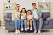 © Studio Romantic - Young happy Caucasian family with two children watching TV together sitting on sofa in living room. Little girl points remote control at camera while sitting next to her mom, brother and dad.