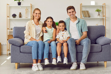 Young happy Caucasian family with two children watching TV together sitting on sofa in living room. Little girl points remote control at camera while sitting next to her mom, brother and dad.