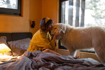 Canvas Print - Young woman cares her huge adorable white dog while lying in bed in tiny bedroom of wooden cabin on nature. House coziness and friendship with pets concept