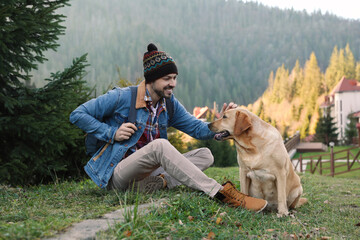 Canvas Print - Happy man and adorable dog sitting on green grass in mountains. Traveling with pet