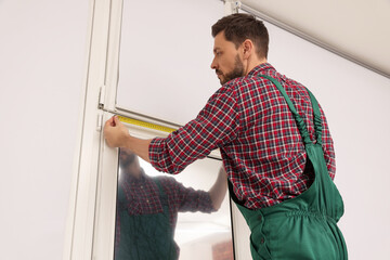 Poster - Worker in uniform using tape measure while installing roller window blind indoors