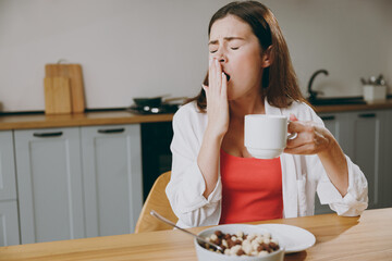 Wall Mural - Young sleepy tired sad european housewife woman wearing casual clothes covering mouth with hand yawn drinking coffee eating breakfast cooking food in light kitchen at home alone. Healthy diet concept.