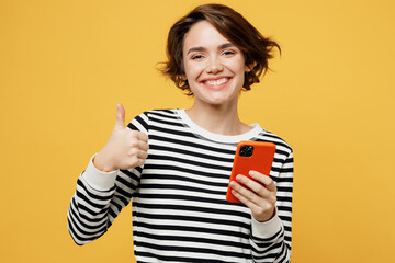 Young fun woman wearing casual striped black and white shirt hold in hand use mobile cell phone in red case show thumb up isolated on plain yellow color background studio portrait. Lifestyle concept.