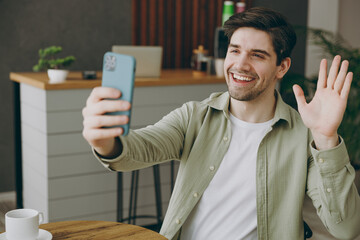 Wall Mural - Young caucasian man wears casual clothes doing selfie shot on mobile cell phone post photo on social network waving hand sits alone at table in coffee shop cafe restaurant indoors rest relax inside.