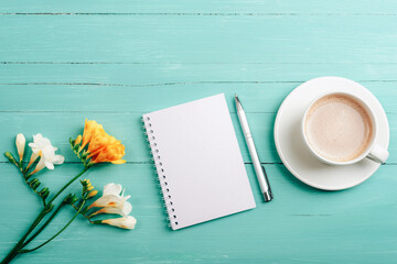 blank notepad, pen and coffee cup on turquoise wooden table with freesia flowers. top view, flat lay