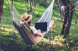 Young woman in a hat using a laptop while lying in a hammock in a summer garden. Summer lifestyle concept, working online
