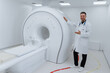 © makedonski2015 - A male doctor in a white coat stands next to a modern MRI machine in the clinic. A smiling man looks into the camera.