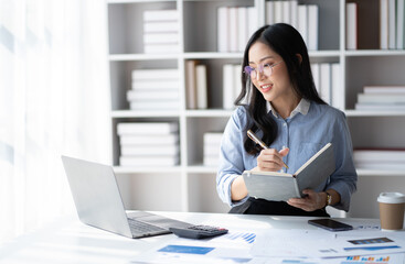 Wall Mural - Young smart Asian businesswoman with glasses writing her idea while working in her office room.