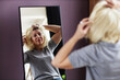 © Seventyfour - Portrait of mature bald woman putting on wig looking in mirror while getting ready in morning