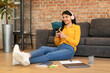 © Prostock-studio - Cheerful teen mexican lady using cellphone wearing headphones texting in social media app, sitting on floor near sofa