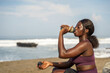 © Jovo Jovanovic/Stocksy - Woman drinking water from a flask sitting at the beach after workout
