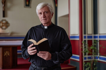 Wall Mural - Portrait of senior priest looking at camera while standing with BIble book in church