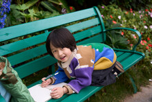 Asian Little Boy Drawing Peacefully On A Bench In A Beautiful Garden