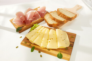 cheese slices with greens and prosciutto meat delicacy on a wooden board. bread is behind. snack on a light background. antipasto under sunlight. dairy and farm products on the table