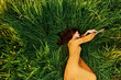 an elegant woman lies in the tall green grass in a long summer orange dress and relaxed posing with her hands enjoying nature and sunny weather with her eyes closed