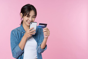 Wall Mural - Young beauty Asian woman shopping payment online with credit card on smartphone and she wearing jean and looking at phone isolated on pink background