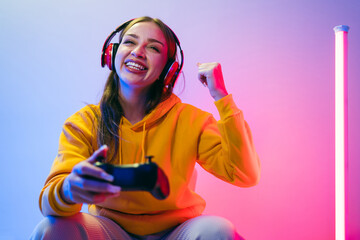 A young Caucasian beautiful woman in a yellow hoodie playing a video game using a joystick and headphones with fun, focus, surprise, and a winning victory face with blue and red neon light.