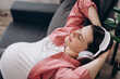 Head shot smiling peaceful young pregnant woman relaxing on comfortable sofa, listening to favorite audio music in modern earphones. Mindful happy future mom enjoying lounge stress free time at home
