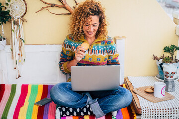 Wall Mural - Cheerful young adult woman using computer sitting and comfy eating cookie. Happy home lifestyle and surfing the net. Modern people work on laptop in leisure online activity. Alternative office
