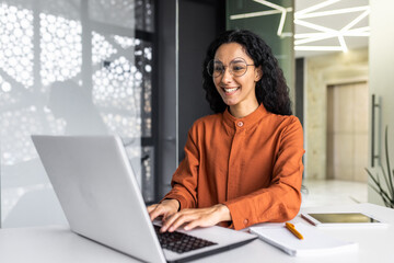 Wall Mural - Happy and smiling hispanic businesswoman typing on laptop, office worker with curly hair and glasses happy with achievement results, at work inside office building