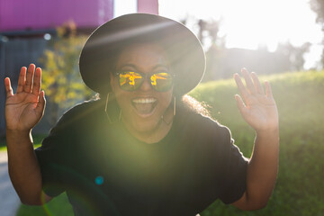 Poster - Happy African american young woman wearing t-shirt laughing looking at camera standing on street. Smiling generation z hipster girl posing outdoor backlit with sunlight portrait