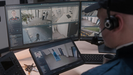 Security officer in headset controls CCTV cameras in office, uses digital tablet and computers. High tech software showing surveillance cameras footage playback on screens. Concept of social safety.