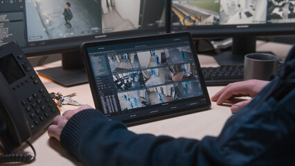 Security operator controls CCTV cameras in office, uses digital tablet and computers with surveillance camera playback on screens. High tech security with facial recognition. Concept of social safety.