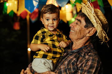 Senior man and his baby grandson celebrating the Brazilian Festa Junina. Portrait of grandfather and his grandson wearing typical clothes and a straw hat during the traditional June festival in Brazil