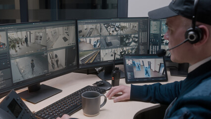 Security officer in headset controls CCTV cameras in office, uses digital tablet and computers. High tech software showing surveillance cameras footage playback on screens. Concept of social safety.
