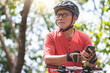 Portrait of Asian Senior Chinese Adult man in helmet holding mobile phone or smartphone while cycling with bicycle at park outdoor. Grandfather exercising with bike riding. Looking away.
