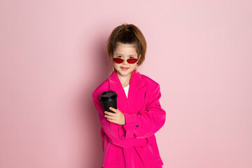 Wall Mural - Portrait of a little girl in an oversized pink blazer and a slim stylish red glasses. Child wearing adult clothes. Isolated on pink background.