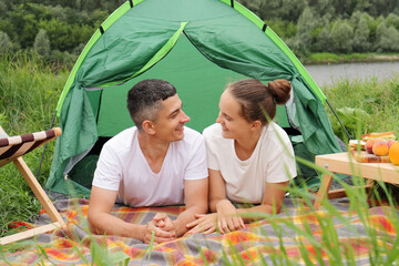 Wall Mural - Joyful couple in nature, woman and man wearing casual clothing laying at tent near the river, happy vacation, romantic weekend, enjoying communication with each other.